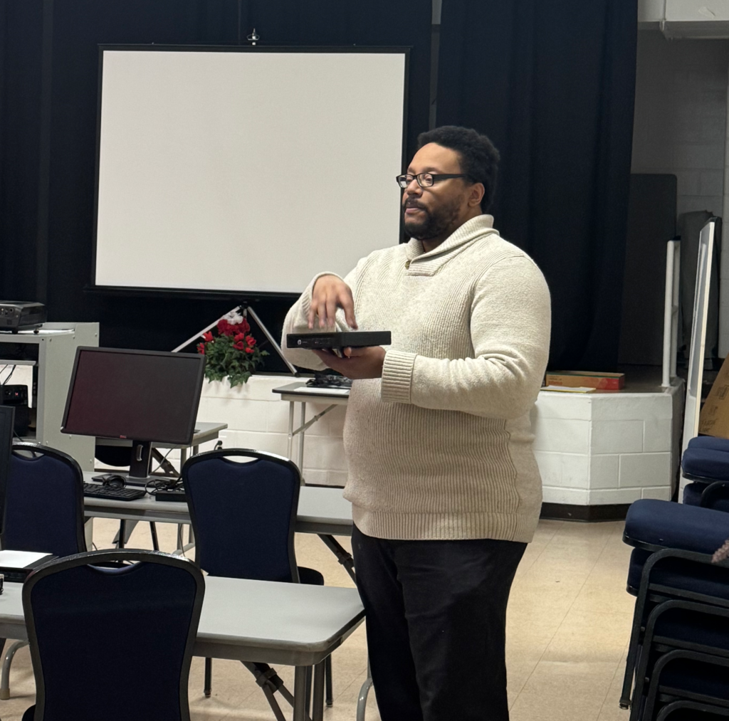 Sean Mitchell, in a white sweater, standing in a classroom holding a mini-desktop computer while instructing the class.